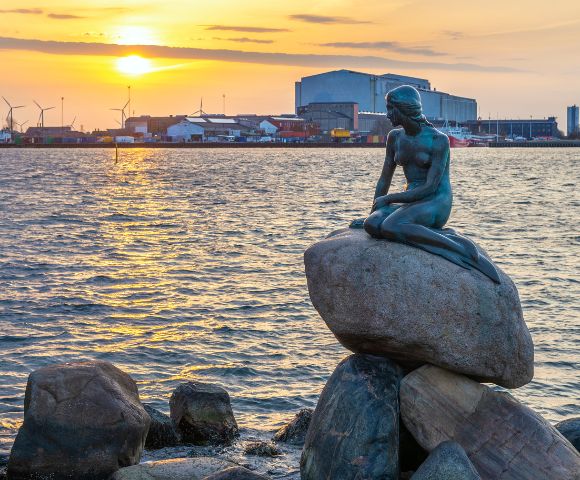 Statue of a mermaid sits on rocks, gazing over a harbor at sunset. Wind turbines and industrial buildings line the distant horizon. Peaceful ambiance.