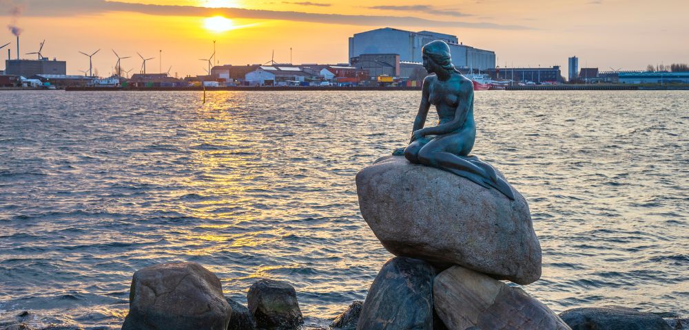 Statue of a mermaid sits on rocks, gazing over a harbor at sunset. Wind turbines and industrial buildings line the distant horizon. Peaceful ambiance.