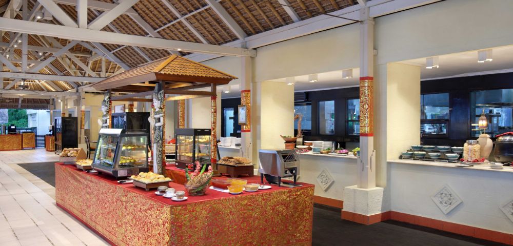 Buffet setup in a spacious open-air dining area with a high thatched roof and white beams. A central table covered in a red and gold ornate cloth displays various food items in glass cases and on plates, with additional food stations along the walls.