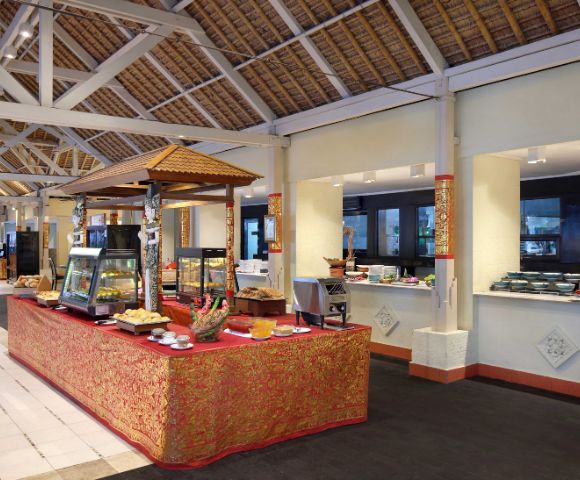 Buffet setup in a spacious open-air dining area with a high thatched roof and white beams. A central table covered in a red and gold ornate cloth displays various food items in glass cases and on plates, with additional food stations along the walls.