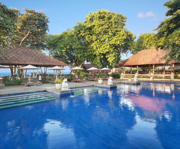 Outdoor swimming pool with clear blue water, surrounded by lush greenery, white statues, and thatched-roof structures under a sunny sky.
