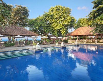 Outdoor swimming pool with clear blue water, surrounded by lush greenery, white statues, and thatched-roof structures under a sunny sky. Outdoor swimming pool with clear blue water, surrounded by lush greenery, white statues, and thatched-roof structures under a sunny sky.