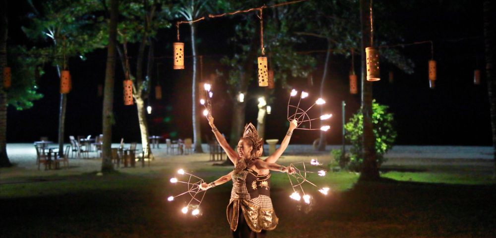 Fire dancer in traditional attire performing at night with multiple torches, surrounded by trees, lanterns, and outdoor seating.