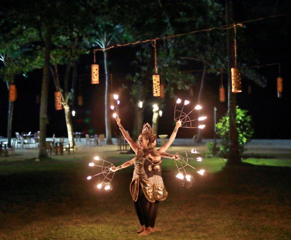 Fire dancer in traditional attire performing at night with multiple torches, surrounded by trees, lanterns, and outdoor seating.