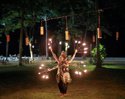 Fire dancer in traditional attire performing at night with multiple torches, surrounded by trees, lanterns, and outdoor seating.