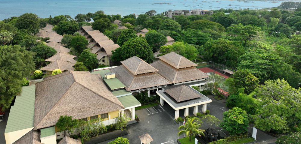 Aerial view of a tropical resort with thatched-roof buildings, lush greenery, connecting pathways, and an ocean in the background with small boats.