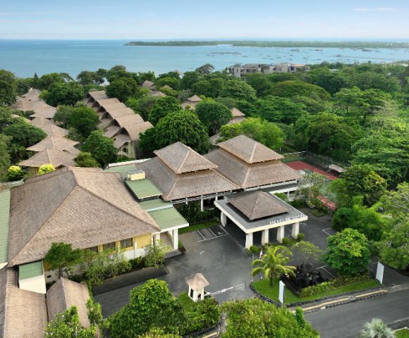 Aerial view of a tropical resort with thatched-roof buildings, lush greenery, connecting pathways, and an ocean in the background with small boats.