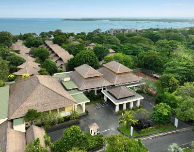 Aerial view of a tropical resort with thatched-roof buildings, lush greenery, connecting pathways, and an ocean in the background with small boats.