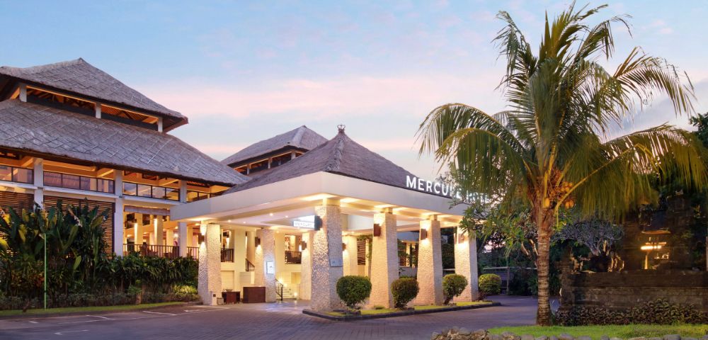 Hotel entrance with stone pillars, warm lighting, and a palm tree, set against a pinkish sky at sunrise or sunset. The building features traditional-style roofing and modern architectural elements.