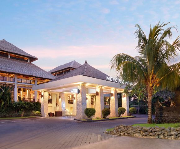Hotel entrance with stone pillars, warm lighting, and a palm tree, set against a pinkish sky at sunrise or sunset. The building features traditional-style roofing and modern architectural elements.