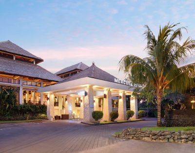 Hotel entrance with stone pillars, warm lighting, and a palm tree, set against a pinkish sky at sunrise or sunset. The building features traditional-style roofing and modern architectural elements.