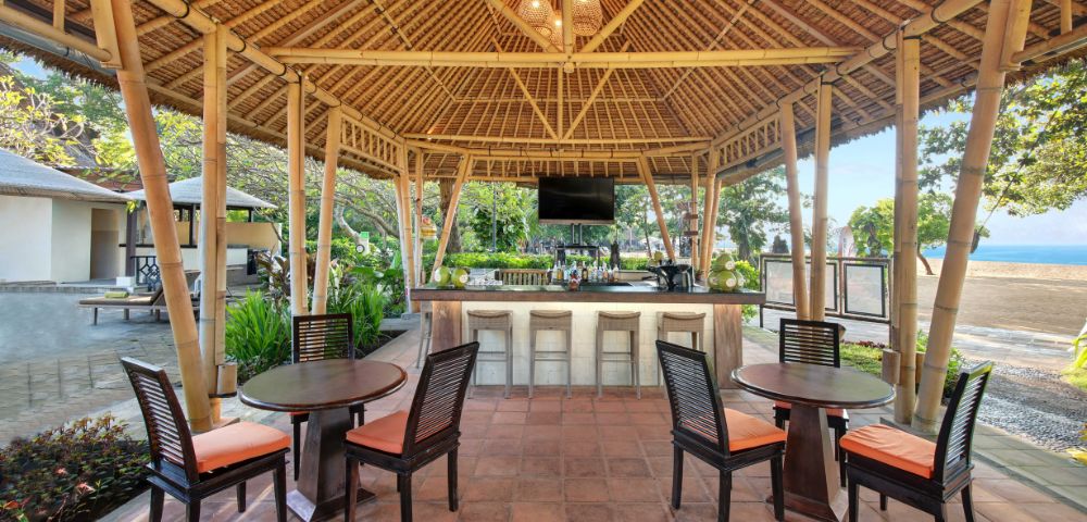 An open-air bamboo pavilion featuring a bar, several tables with chairs, and a view of greenery and a beach in the background.