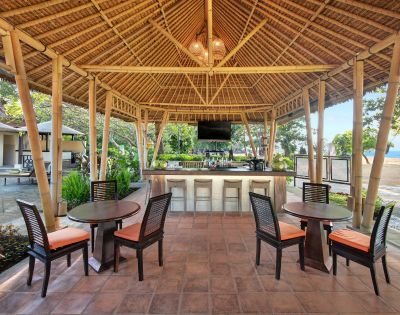 An open-air bamboo pavilion featuring a bar, several tables with chairs, and a view of greenery and a beach in the background.