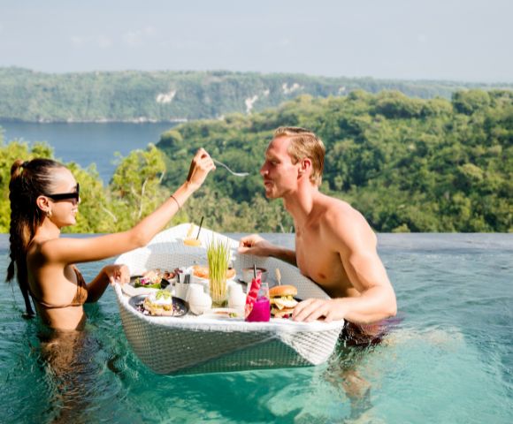 A couple enjoys a floating breakfast in an infinity pool overlooking lush hills. The woman feeds the man amid a cheerful, relaxed setting.