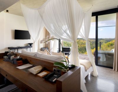 Elegant hotel room with a canopy bed draped in sheer white fabric, wooden desk with orchid, TV on wall, and large window showcasing scenic hilly view.