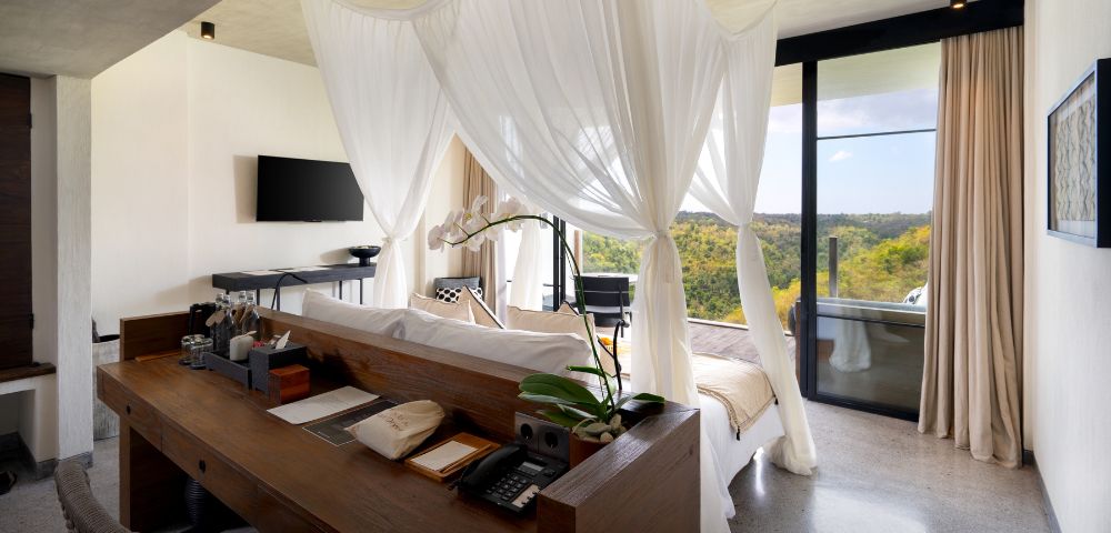 Elegant hotel room with a canopy bed draped in sheer white fabric, wooden desk with orchid, TV on wall, and large window showcasing scenic hilly view.
