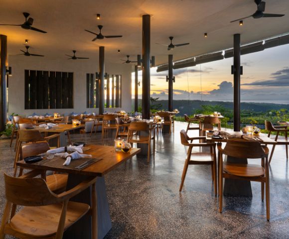 Elegant, open-air restaurant at sunset, with wooden tables and chairs, ceiling fans, warm lighting, and large windows showcasing a scenic horizon.