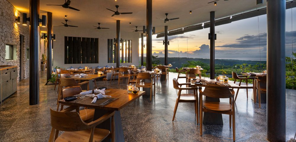 Elegant, open-air restaurant at sunset, with wooden tables and chairs, ceiling fans, warm lighting, and large windows showcasing a scenic horizon.