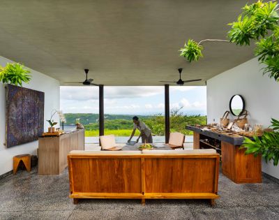 Open-air room with wooden furniture and greenery. A person sets a table with a scenic view of lush hills and a blue sky in the background. Calm ambiance.
