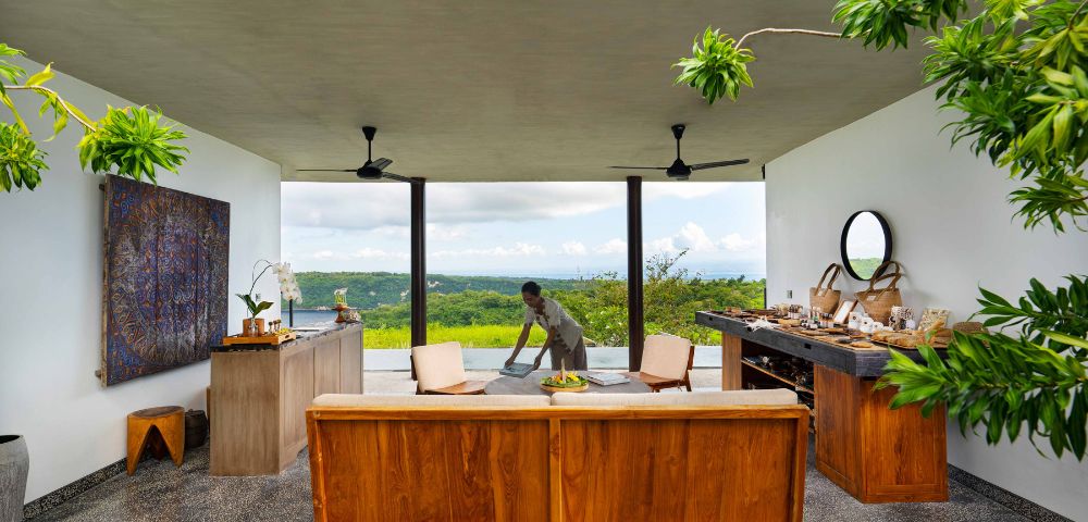 Open-air room with wooden furniture and greenery. A person sets a table with a scenic view of lush hills and a blue sky in the background. Calm ambiance.