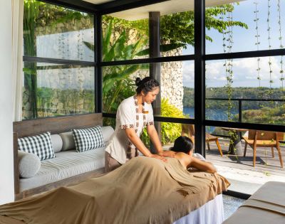 A serene spa scene with a therapist giving a massage in a sunlit room. Large windows reveal lush greenery and a tranquil landscape, evoking relaxation.