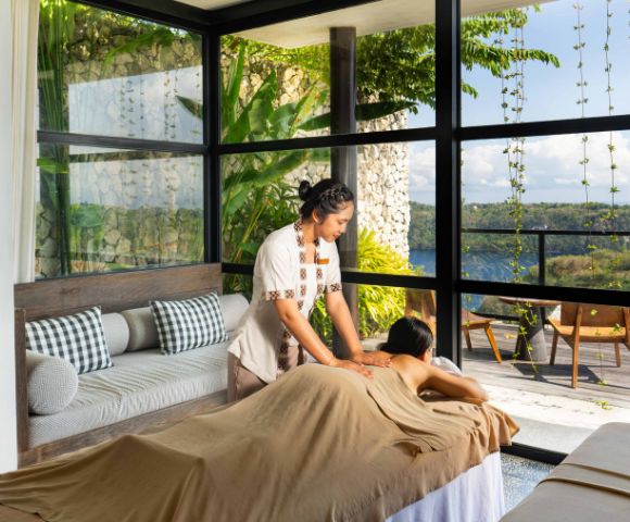 A serene spa scene with a therapist giving a massage in a sunlit room. Large windows reveal lush greenery and a tranquil landscape, evoking relaxation.