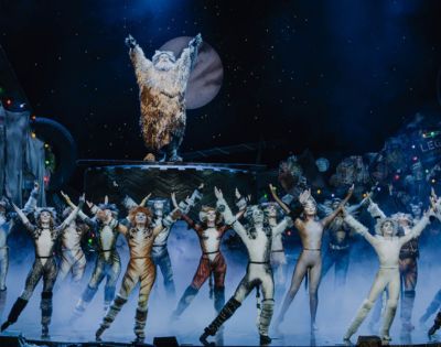 A theatrical stage scene with performers in cat costumes, arms raised, under blue lights. A figure in a shaggy outfit stands elevated, framed by a moon backdrop.