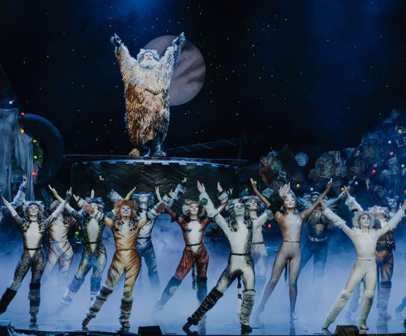 A theatrical stage scene with performers in cat costumes, arms raised, under blue lights. A figure in a shaggy outfit stands elevated, framed by a moon backdrop.
