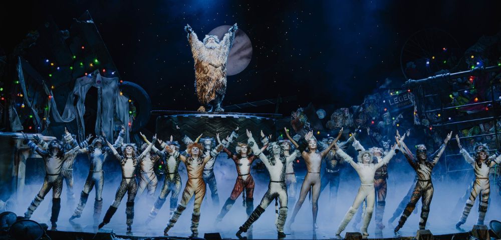 A theatrical stage scene with performers in cat costumes, arms raised, under blue lights. A figure in a shaggy outfit stands elevated, framed by a moon backdrop.