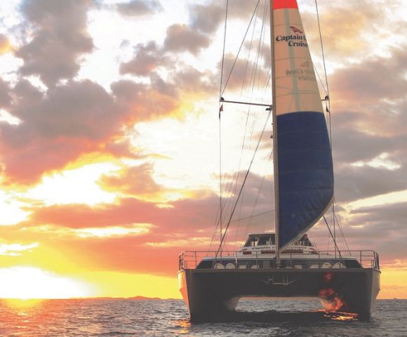 A catamaran sails towards the viewer on a calm ocean during a vibrant sunset. The sky is filled with dramatic clouds and orange, pink, and yellow hues.