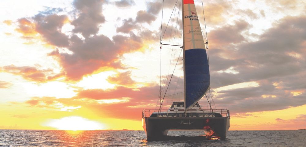 A catamaran sails towards the viewer on a calm ocean during a vibrant sunset. The sky is filled with dramatic clouds and orange, pink, and yellow hues.