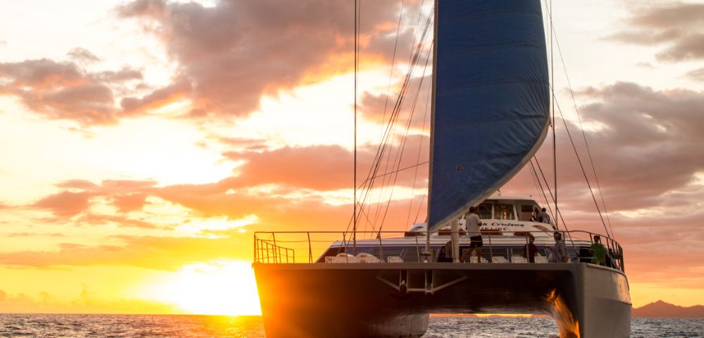A catamaran with red, white, and blue sails glides on the ocean at sunset. The vibrant sky and serene water create a peaceful, scenic atmosphere.