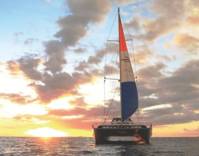 A catamaran sails towards the viewer on a calm ocean during a vibrant sunset. The sky is filled with dramatic clouds and orange, pink, and yellow hues.