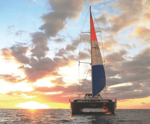 A catamaran sails towards the viewer on a calm ocean during a vibrant sunset. The sky is filled with dramatic clouds and orange, pink, and yellow hues.