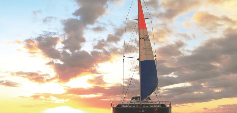 A catamaran sails towards the viewer on a calm ocean during a vibrant sunset. The sky is filled with dramatic clouds and orange, pink, and yellow hues.