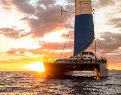 A catamaran with a tall red, white, and blue sail glides on the ocean during a vibrant sunset, casting a serene and adventurous mood.