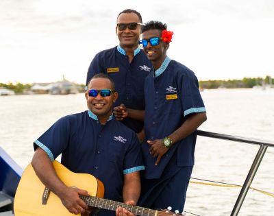 Three men wearing blue uniforms and sunglasses stand on a boat. One holds a guitar, another wears a flower behind his ear. They all smile warmly, suggesting a cheerful atmosphere.