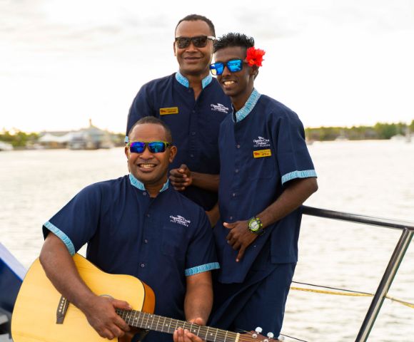 Three men wearing blue uniforms and sunglasses stand on a boat. One holds a guitar, another wears a flower behind his ear. They all smile warmly, suggesting a cheerful atmosphere.