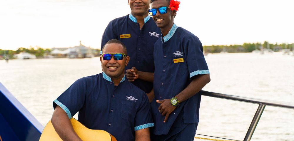 Three men wearing blue uniforms and sunglasses stand on a boat. One holds a guitar, another wears a flower behind his ear. They all smile warmly, suggesting a cheerful atmosphere.