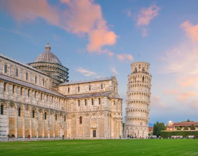 The image shows the Leaning Tower of Pisa at sunset, with a vibrant sky. Surrounding buildings and green lawn create a serene and historic atmosphere.