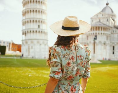 Woman in a floral dress and straw hat, facing the Leaning Tower of Pisa on a sunny day. The scene conveys a sense of wanderlust and exploration.