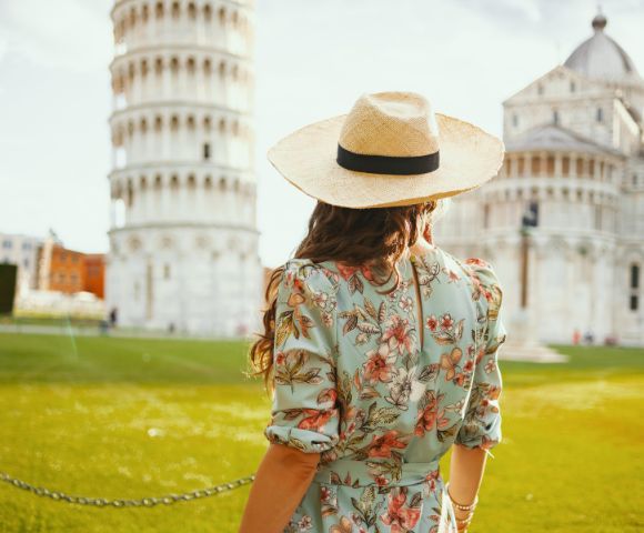 Woman in a floral dress and straw hat, facing the Leaning Tower of Pisa on a sunny day. The scene conveys a sense of wanderlust and exploration.
