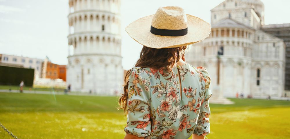 Woman in a floral dress and straw hat, facing the Leaning Tower of Pisa on a sunny day. The scene conveys a sense of wanderlust and exploration.