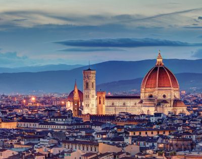 A panoramic view of Florence at dusk, featuring the illuminated Duomo and bell tower. The skyline is set against rolling hills under a twilight sky.
