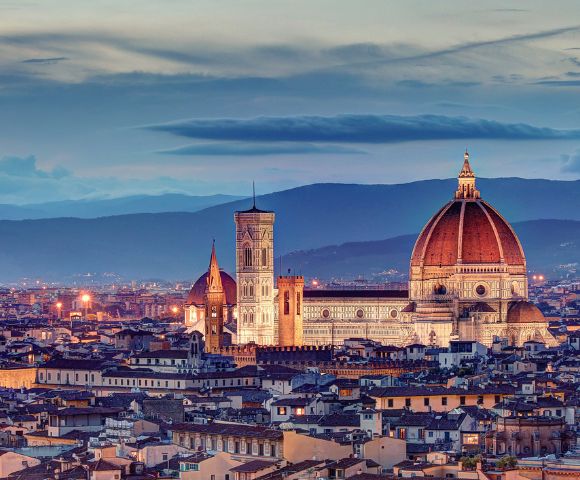A panoramic view of Florence at dusk, featuring the illuminated Duomo and bell tower. The skyline is set against rolling hills under a twilight sky.