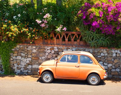 An orange vintage car is parked by a stone wall with lush greenery and vibrant pink flowers. The scene is sunny, evoking a cheerful, nostalgic tone.