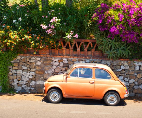 An orange vintage car is parked by a stone wall with lush greenery and vibrant pink flowers. The scene is sunny, evoking a cheerful, nostalgic tone.