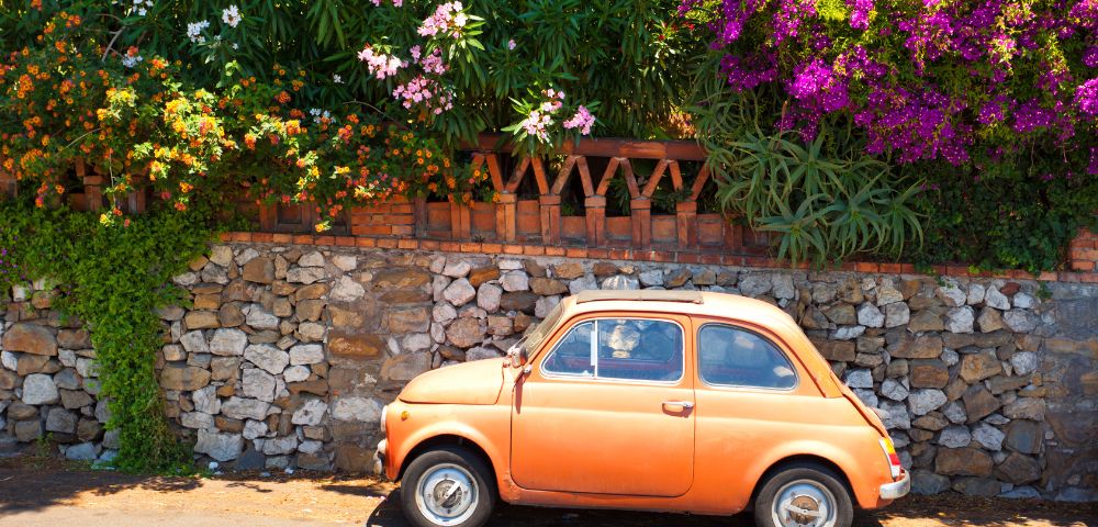 An orange vintage car is parked by a stone wall with lush greenery and vibrant pink flowers. The scene is sunny, evoking a cheerful, nostalgic tone.