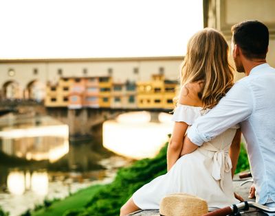 A couple sits closely, overlooking a river and historic bridge at sunset. They exude a romantic and peaceful vibe. A straw hat rests beside them.