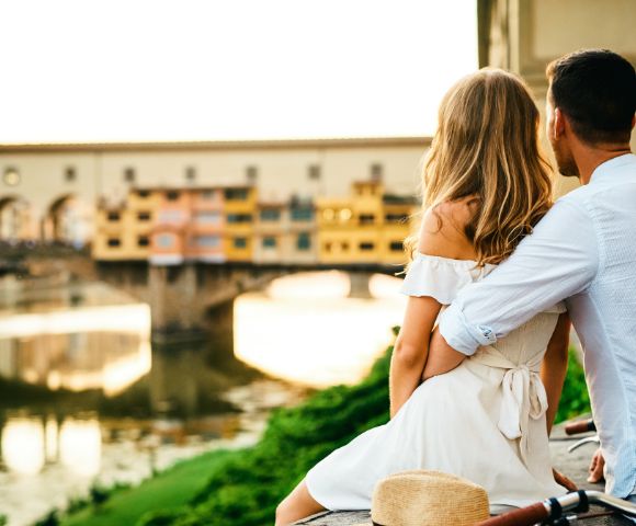 A couple sits closely, overlooking a river and historic bridge at sunset. They exude a romantic and peaceful vibe. A straw hat rests beside them.